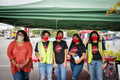 5 volunteers standing under a green tent in Black El Pueblo t-shirts wearing red masks. In spanish. 5 voluntarios parados debajo de una carpa verde con camisetas negras de El Pueblo y máscaras rojas. En español.